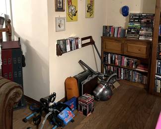 In this photo, there are a number of different items, including a stationary cycling machine, a vacuum cleaner and accessories, a few pieces of art on the wall above DVDs or Blu-Rays.