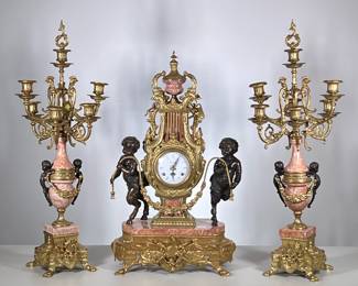 (3pc) Italian Imperial Clock Garniture | Intricate gilt metal and pink marble three piece clock garniture, comprising a large lyre and urn form mantel clock with double rooster crest, the clock flanked by two bronze satyre figures, the clock face marked "Imperial" and having Roman numeral markers, the movement marked Franz Hermle; centered by two 7-arm urn-form candelabras decorated with gilt garland and with bronze putti-form handles. -  h. 23.5 x w. 14.5 x d. 8.5 in
