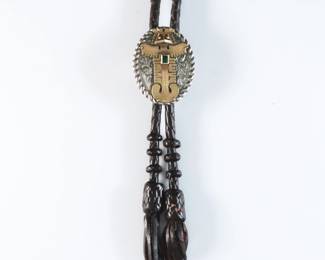 A vintage Pre Columbian style 18k Gold and Silverplate bolo tie. Features a Silverplate clasp with an 18k Gold figure and Green gemstone accent on a braided leather tie. Clip marked "18k" and "Spiros" for Spiros Inc. Jewelry, Fort Lauderdale, FL. Minor wear to patina and surface. Clasp is 1 1/2" high, 20" long overall.