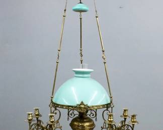 A late 20th century kerosene chandelier. Brass body with cast mask details features a Green cased glass shade and matching canopy over an embossed font surrounded by three scrolling arms with four candle holders each. Some wear. 54" high plus pull down.