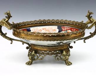 A Japanese Imari mounted center bowl. Low porcelain bowl with polychrome scenic and floral decoration in a Bronze mount with two Dragon handles and paw feet. Frame and handle with soldered adjustments, porcelain with some surface wear. 17 1/2 x 8 1/2" high overall.