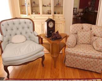 A vintage French style carved arm chair in button back blue fabric, a china display cabinet, a triangular 2-tier side table, an antique Gilbert Manufacturing American mantel shelf clock, an upholstered skirted armchair