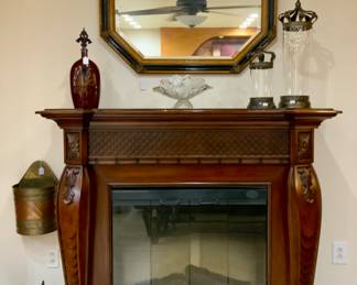 Elongated Black & Gold Wood Frame Bevel (42.5” x 29.5”)Mirror above a Vintage Empire Design Cherry Fireplace Mantle Flanked with Cabriole Style Legs with Shell and other carvings (60”W x 22”D x 48”H). Ruby Red Fleur de Lis Bottle, Crystal Clear Inc. “Moravia” Pedestal Banana Bowl and 2 Graduated Cylindrical Canisters on Metal Base and Metal CrownLids w/ Cross Finial