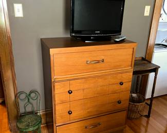 A Mengel Chest of Drawers, a Sylvaniaflat screen TV on top, and on the left is a child's spectacle eye glass back ice cream chair, and on the right - an end table, a round mirror above.