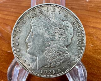 This lot features a 1921 Morgan Dollar. The Morgan silver dollar, designed by Mint Chief Engraver George T. Morgan and produced from 1878 to 1904, with a brief reissue in 1921, was a significant part of American coinage. It was minted as a standard silver dollar after the Coinage Act of 1873 ended the free coining of silver. The Morgan dollar, known for its size and weight, became a popular coin, especially in the American West, and is now a favorite among collectors.