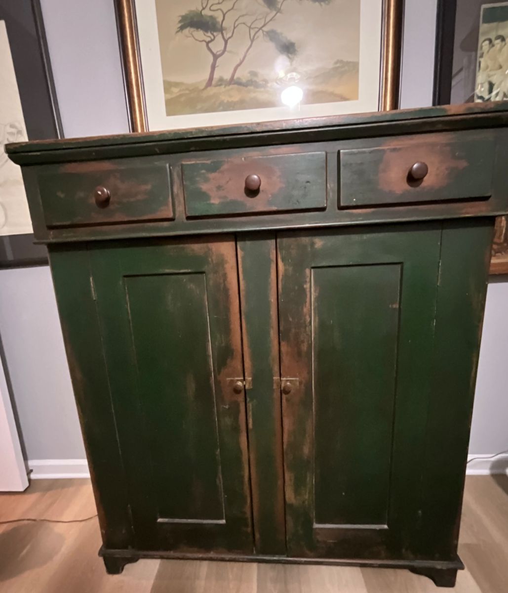 antique jelly cupboard, likely dating from the mid-19th century. It features a simple, utilitarian design, common in rural American furniture of the period. The cupboard is constructed from wood, probably pine or poplar, and is painted in a dark green color with a distressed finish, revealing hints of the original wood underneath. It has three drawers above two doors, and simple wooden knobs.