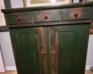 antique jelly cupboard, likely dating from the mid-19th century. It features a simple, utilitarian design, common in rural American furniture of the period. The cupboard is constructed from wood, probably pine or poplar, and is painted in a dark green color with a distressed finish, revealing hints of the original wood underneath. It has three drawers above two doors, and simple wooden knobs.