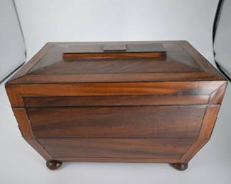 Elegant and finely crafted, this early 19th-century tea chest is a beautiful example of Regency-period design. Made from richly grained mahogany with delicate boxwood or satinwood inlay, the chest features faceted sides and a raised lid, all resting gracefully on turned bun feet.
The interior is lined in red fabric and fitted with two original lidded compartments for loose tea storage, each adorned with small bone or ivory knobs. At center sits a cut glass mixing bowl, scalloped and patterned, accompanied by a vintage silver-plated tea caddy spoon—used to blend teas before serving, as was customary in refined households of the time.
Both functional and decorative, this tea chest reflects the era’s deep appreciation for ritual and craftsmanship. A charming and rare find, ideal for collectors of Georgian and Regency antiques, or as a statement piece for a traditional home. Estimated date: 1810–1820