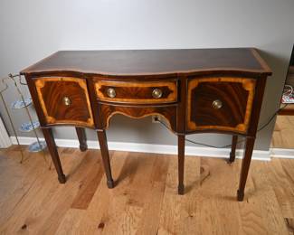 Elegant Federal-style bow front side table, with gorgeous flame mahogany veneer and contrasting inlay, all wrapped in a graceful curved silhouette. Two drawers offer practical storage, each fitted with traditional round brass knobs. Tapered legs complete the neoclassical look.