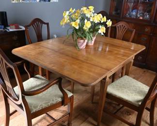 A timeless dining set featuring a classic drop-leaf table with tapered, fluted legs and brass-capped feet, paired with six elegant Hepplewhite-style chairs. The chairs feature shield backs and updated upholstered seats in a tasteful geometric pattern.