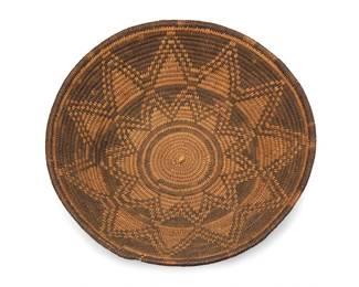 306
An Apache basketry tray
Early 20th century; Southwest United States
A large coiled low bowl with two floral starburst motifs to center
3.375" H x 12" Dia.
Estimate: $400 - $600