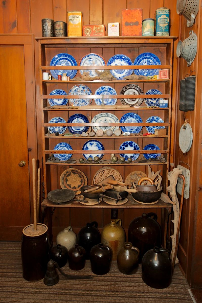 Vintage tins and boxes; shelving unit with flow blue china; Antique and vintage cast iron cooking pans, vintage primitive carved kitchen tools and household tools, a collection of antique pottery jugs; vintage primitive wrought iron skillets, griddle and three-legged long handled pot; vintage Native American woven bowl/basket