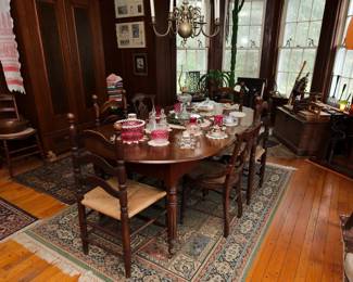 Beautiful extendable wooden dining table shown with some of the antique ladder back and cane-seat chairs available; beautiful area rugs, a tall two-door wardrobe, a selection of antique porcelain dishware, cranberry cut to clear glass, vintage crystal knife rests, framed antique photos, and more.