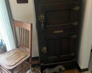 Antique wood icebox with brass hardware and original metal nameplate.
Vintage oak slat-back chair with distressed seat.
Large stoneware crock jug with handle.
Cast dog sculpture in resting position.