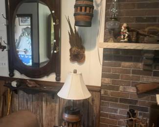 Large oval wood-framed wall mirror, hanging wooden barrel decor, pheasant feathers mounted on plaque, rustic wood shelf with small antiques, vintage oil lamp, ceramic elk figurine, barrel-themed table lamp on fur-covered stand.