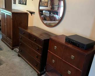 wood armoire with paneled doors and lower drawer.
Dark wood four-drawer dresser with metal knobs.
Mid-century modern style record cabinet with brass square pulls and open lower shelf.
Round wall mirror with wood frame.
Wall-mounted corner display shelves.