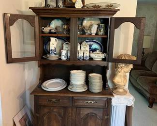 Dark wood china cabinet with glass doors, drawers, and lower storage.
Large set of decorative plates, teacups, and ceramic pitchers with blue and white patterns.
Antique pendulum wall clock with roman numerals.
Victorian porcelain doll with lace dress.
Decorative bust statue on white pedestal column.
Framed and frameless wall art (various sizes).