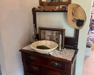 Antique wooden washstand with ornate drawer pulls.
Decorative ceramic wash basin with floral design.
Vintage oil lamp with clear chimney.
Straw sunhat with floral accent.