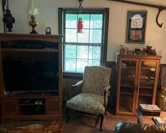 Wood entertainment center. flat-screen TV and lower media.
Vintage oil lamps (amber, red, and clear glass) on display and hanging.
Upholstered armchair with carved wood frame and floral damask fabric.
Glass-front curio cabinet with wood frame and dual doors.
Wall-mounted antique cant hook.
Framed clock print and rustic miniature tractor and automobile decor.
Sheepskin-style area rug.