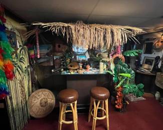tiki bar setup and accessories, with two rattan and leather bar stools, straw thatch roof canopy, tropical decorations including palm leaves, colorful leis, framed artwork, and various collectibles on and behind the bar. patterned area rug.