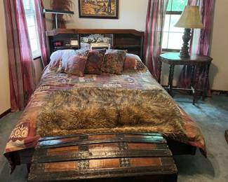Rustic queen-size bed with built-in headboard storage and southwestern bedding set.
Large curved-top steamer trunk with black metal trim and wood slats.
Round carved wood side table with pedestal base.
Decorative table lamp with fabric shade.
