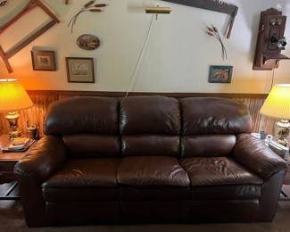Brown leather sofa with plush cushions and wide armrests.
Matching brass table lamps with pleated shades.
Wood end tables with drawers and brass hardware.
Vintage crank wall phone mounted on wood.
Rustic hand saw and other antiques displayed as wall decor.
Framed wildlife and landscape prints.