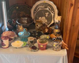 This table is on the right side of the living room close to the dining table. It features three vintage trays, several pieces of pottery, a blue candy dish, an old rolling pin, and a set of rooster and hen figurines. There is also a bird carved from a coconut shell.