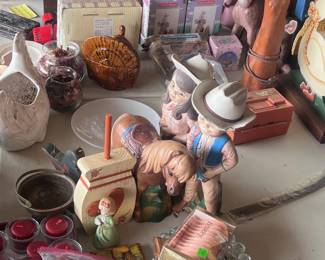This table is located in the garage. The tables in the garage will all have a wide range of things on each table. Some items you probably will have no idea what they are for. This table has several small candles, a vintage Cypress lamp, white rooster planter, and lots of other odds and ins. The horse and cowgirl figures are in real good condition but the cowboy has damage.