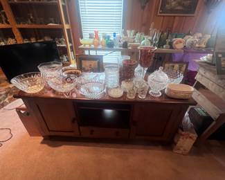 This entertainment center is located in the center of the living room just in front of the fireplace. The entertainment center has cabinets on both ends and a drawer in the bottom middle. There is a variety of lead crystal pieces, bowls, candy dishes, and other pieces on top of the entertainment center. A wooden magazine holder is on the floor to the left.