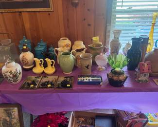 This table is located on the right side of the living room. It has lot of collectable items. There is a green McCoy vase along with two blue canaster sets, several very old pitchers, a clay chicken waterer (very old), and three Japanese hand painted pictures. In addition there is a handblown bottle with removable cap and a vintage thermos bottle. Many items on the floor including an old wall picture.