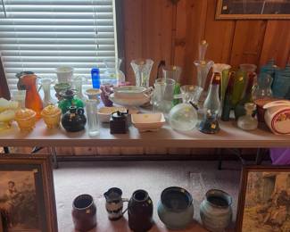 This table is located in on the right side of the living room. The table has a wide range of different vintage colored glass, a clear glass tea kittle, two white with pink rimmed vases, a green colored oil lamp, and several pieces of pottery. On the floor there are two small churns, two large pottery urns, and a pottery pitcher. More very pretty wall pictures are located here as well. There are things still hanging on the wall over these tables that are priced.
