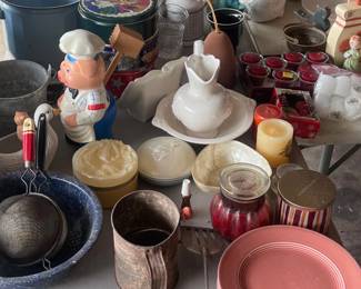 This table is located in the garage. It houses lot of vintage collectable pieces including a flower shifter, vintage sieves, blue porcelain slop bucket, white pitcher and basin, very old eye glass in the black cases.