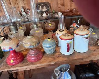 These items are located in the center of the living room. This table has several vintage oil lamps of at least two different types, three coffee pots, a stack of old platters of different shapes, sizes, and patterns, along with other porcelain and glass items. On the floor there is a large water basin and wash bowl along with a small chest.