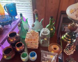 This table is located on the right side of the living room. It features a vintage cookie jar, several handblown green wine decanter jugs. There is also a top to a #3 churn and a zip lock bag of old match books.