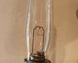 Aladdin 23 Kerosene Oil Lamp. The base of the lamp is made of white glass with a painted floral design featuring small flowers. The burner assembly, located between the glass chimney and the white glass base, is made of brass with an intricate ornate design.