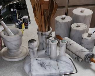 Elegant and cohesive kitchen collection of white marble with gray veining. Pictured: round cheese cutting board with, salt and pepper shaker sets, a large mortar and pestle, four-piece canister set, a rolling pin. a utensil
holder filled with an assortment of wooden utensils, including spoons, spatulas, and forks, large and medium lazy susans.
