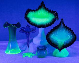 Seen in blacklight: An early art glass vase, Rubina Verde uranium glass possible Hobbs Brockunier; antique cranberry glass vase with pulled petals flared at the rim and applied light green uranium glass foot; two 19th century opalescent and custard cased uranium glass Jack in the Pulpit art glass vases with double crimped ruffled rims