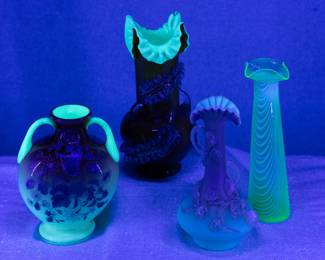 And here they are under UV light source: A Harrach Glassworks peachblow art glass vase of twin handle moon flask form, in cased uranium glass with enameled butterfly & blossom decoration; a Stevens & Williams art glass vase of cased orange and white uranium glass with applied clear rigaree feather-like decoration and ruffled crimped rim, a beautiful antique Stevens & Williams cased opaque matt finish uranium glass vase, shading from pale ivory to warm cinnamon, with applied flowering vine decoration frosted rigaree glass; and a Nailsea style opalescent pulled drapery design uranium glass vase