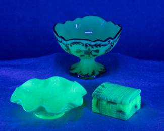 Under black light: EAPG - A Northwood Intaglio pattern custard glass UV reactive berry glass, a Northwood uranium custard glass "Pansy" pattern ruffled edge bowl, and an Akro Agate Company No. 804 Treasure Trunk dusting powder jar, uranium slag glass.