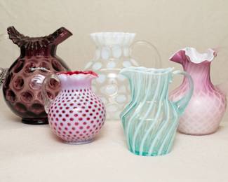 Five different 20th century art glass pitchers, three are by Fenton – a Fenton cranberry and opalescent glass pitcher with clear handle, an L. G. Wright Glass Thumbprint Amethyst pitcher with clear handle, a Fenton white coin dot clear glass pitcher, a Fenton opalescent spiral optic teal pitcher, and a Mt. Washington Glass pink cased diamond quilted MOP satin glass pitcher