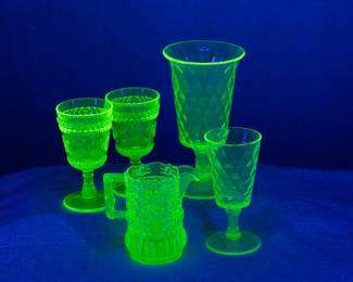 Seen under UV light: A pair of L G Wright Glass Wildflower vaseline glass stemmed glasses, an EAPG 1890's Bryce Bros Vaseline Diamond Quilted uranium glass stemmed vase and smaller goblet, and an EAPG vaseline glass star dot quilt pattern creamer or syrup jug.