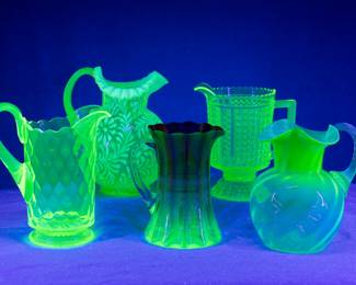 And under black light, see the uranium glass glow! Blacklight photo of the vintage and antique uranium glass or vaseline glass pitchers, including a Fenton glass Daisy and Fern pattern pitcher, a Rubina Verde glass pitcher with enamel and gilt decoration, a hobnail pattern pitcher and a Fenton Glass uranium glass pitcher with flared rim and applied clear handle