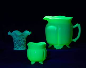 Under black light: Uranium glass including an antique Northwood custard glass Grape Arbor pattern vase, and a Tarentum Glass Co Georgia Gem pattern jadeite creamer 