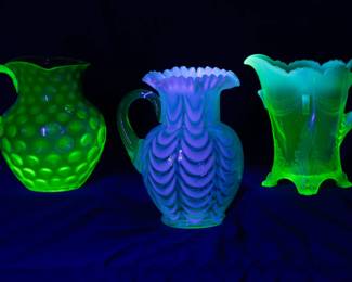 Under black light, another view of the three uranium glass pitchers in Rubina glass, pulled feather patter, and opaline glass