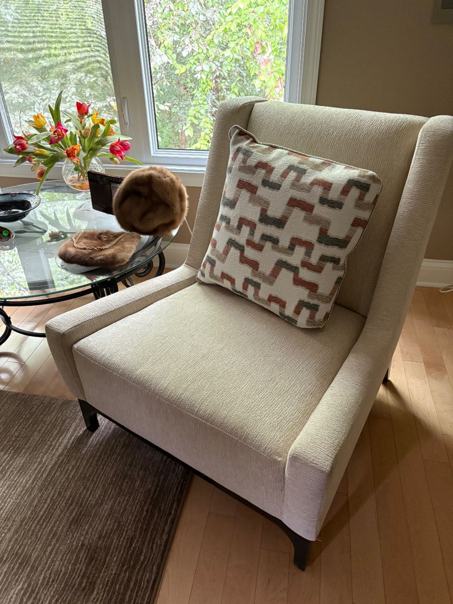 Oversized occasional chair (two available) and view of a glass round low-profile metal base cocktail/coffee table, and mink hat and purse.