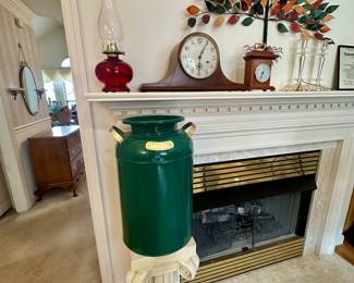 Green butter churn displayed on classic pedestal in front of fireplace, complemented by vintage mantel decor and colorful metal tree wall art.
