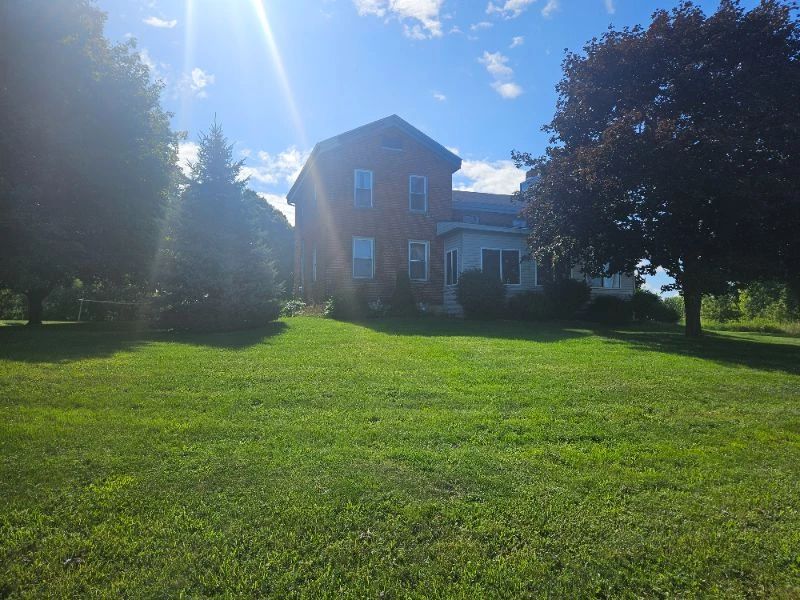 Exterior view of the property featuring a two-story brick house with large windows, a neatly trimmed front lawn, and mature trees under bright sunshine.