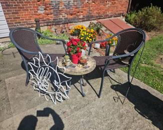 Outdoor bistro set featuring two brown wicker chairs and a round stone-top table; includes a decorative metal sunburst wall art leaning against one chair and a pot of bright flowers on the table.