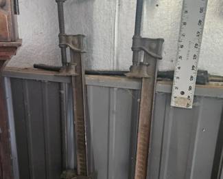 Collection of long‑handled yard tools including shovels, pitchforks and a yardstick ruler leaning against a metal shed wall.