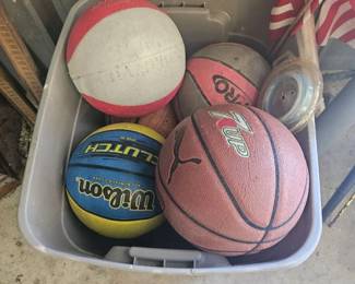 Plastic tub filled with sports balls including basketballs, a soccer ball and baseballs, with an American flag on a stick leaning behind.